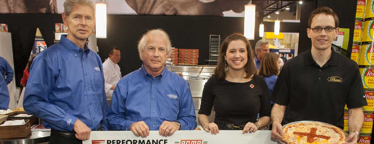 Four people stand behind a counter at a promotional event, with one man holding a pizza. Stacks of canned tomato products and event signage are visible in the background.